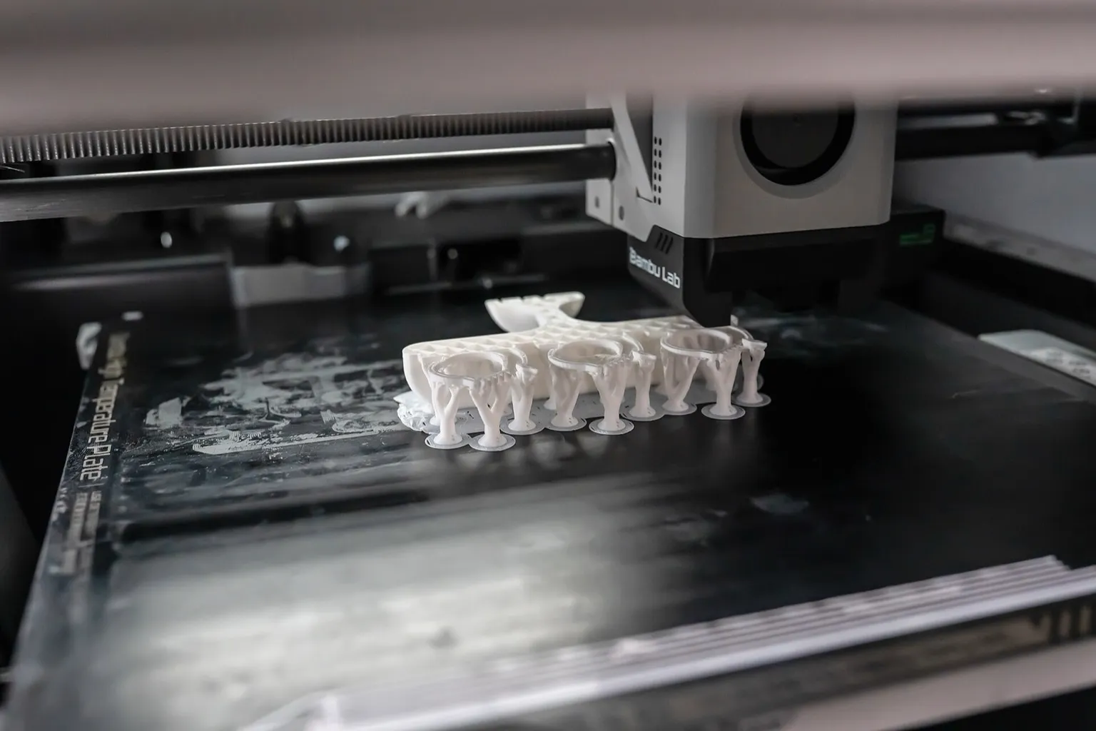 A Bambu Lab toolhead printing a white part on tree supports over a dark Bambu High Temperature Plate, with the plate's printed name along its left edge and white residue on the surface