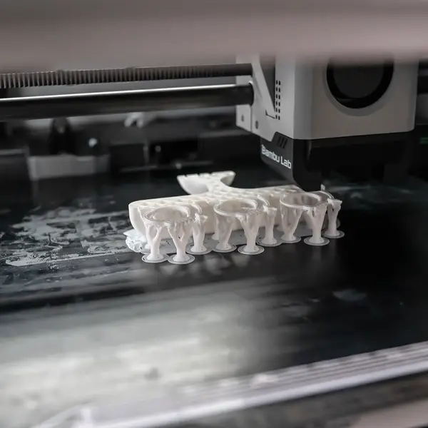 A Bambu Lab toolhead printing a white part on tree supports over a dark Bambu High Temperature Plate, with the plate's printed name along its left edge and white residue on the surface